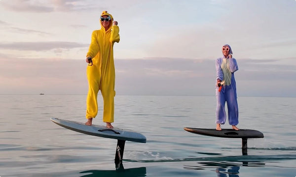Two people in colorful onesies standing on electric hydrofoil boards on a calm body of water.